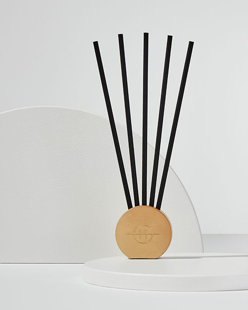 A minimalist gold stand with a circular base and five thin, elongated black stems protruding vertically from the center. The stand is placed against a white background with a subtle shadow cast on a curved white panel behind it, suggesting an elegant and modern design aesthetic.