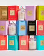 An array of glass candles, each paired with its respective packaging box. The boxes come in a variety of vibrant colors including pink, yellow, blue, red, and purple. Each box has a unique name printed on it such as “Kyoto in Bloom,” “One Night in Rio,” “Arabian Nights,” and others, suggesting different scents inspired by various locations around the world.