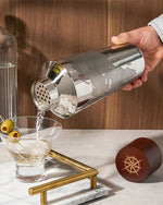 A hand pours liquid from a shiny cocktail shaker into a martini glass with olives on a toothpick. The scene is set on a marble counter, exuding elegance.