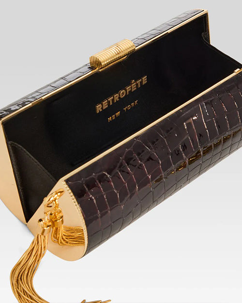 An open cylindrical crocodile-skin clutch with gold sides, a cylindrical gold ribbed clasp and tassel on the side. The interior of the bag is black, and the brand name "RETROFÊTE" along with "NEW YORK" is printed in gold letters inside the bag. The background is white.