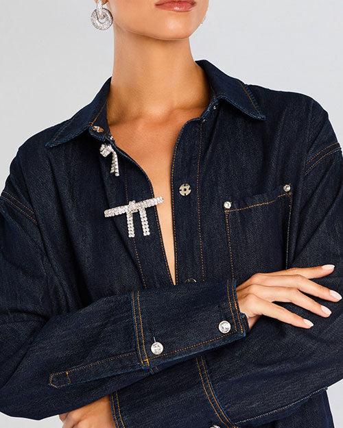 The image displays the torso of a person wearing a dark denim shirt with distinctive silver embellishments. The shirt features silver buttons, a chest pocket. The person’s face is not visible, and their arms are crossed.