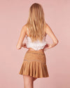 Model showing the back of brown skirt. Model wearing a white lace top and a brown pleated skirt, posing against a pink background