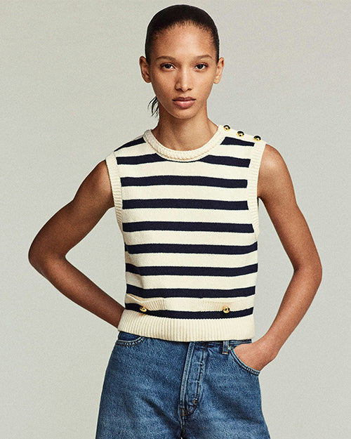 A model is wearing a sleeveless, knitted top with horizontal black- and cream-colored stripes. The top has a round neckline and features 3 gold-colored buttons on the left shoulder and 2 near the waistline. The person is also wearing high-waisted blue jeans with hands placed on hips. The background is plain.