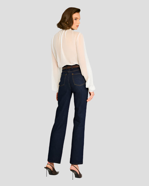 The backside view of a person standing against a plain background, wearing a sheer white blouse with long sleeves. The blouse is tucked into high-waisted dark blue jeans, which are secured with a brown belt featuring a gold buckle. The person is also wearing black heels.