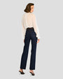 The backside view of a person standing against a plain background, wearing a sheer white blouse with long sleeves. The blouse is tucked into high-waisted dark blue jeans, which are secured with a brown belt featuring a gold buckle. The person is also wearing black heels.