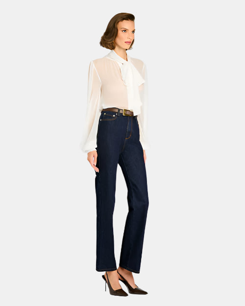 A person is standing against a plain background, wearing a sheer white blouse with long sleeves and a large bow tied at the neck. The blouse is tucked into high-waisted dark blue jeans, which are secured with a brown belt featuring a gold buckle.