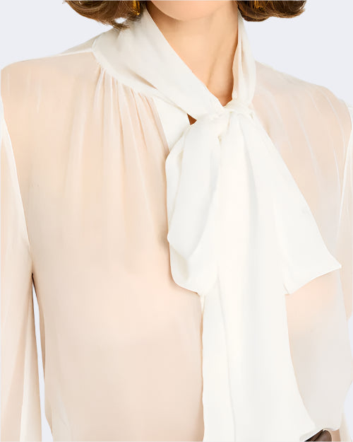 A close-up frontal view of a person wearing a sheer white blouse with a large bow tied at the neck. The tie has a fuller white color.