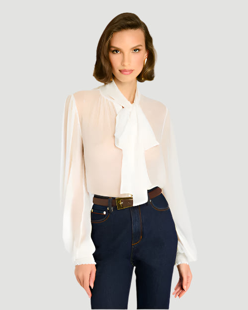 A person is standing against a plain background, wearing a sheer white blouse with long sleeves and a large bow tied at the neck. The blouse is tucked into high-waisted dark blue jeans, which are secured with a brown belt featuring a gold buckle.
