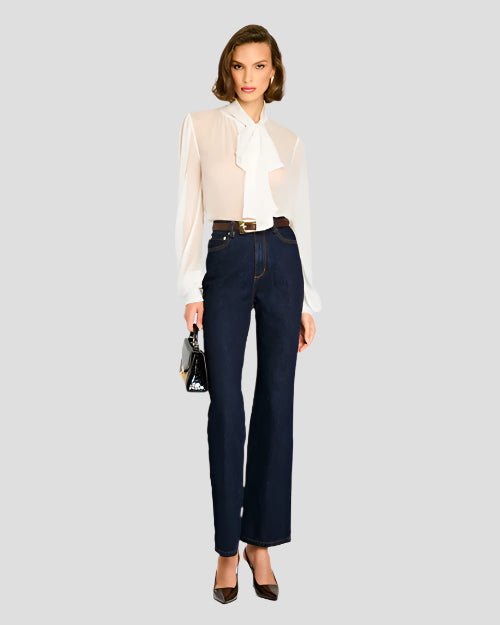 A person is standing against a plain background, wearing a sheer white blouse with long sleeves and a large bow tied at the neck. The blouse is tucked into high-waisted dark blue jeans, which are secured with a brown belt featuring a gold buckle. The person is also holding a small black bag.