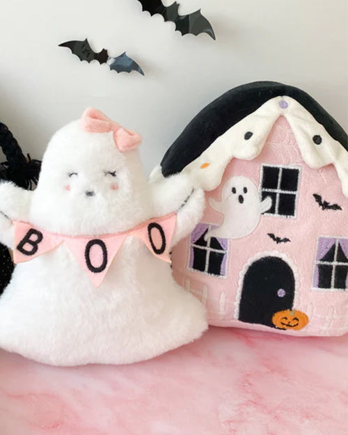 A plush white ghost with a pink bow and a 'BOO' banner stands beside a pink plush haunted house featuring black windows, a black door, small bats, a ghost, and an orange pumpkin near the entrance. Bat decorations are visible on the wall in the background.
