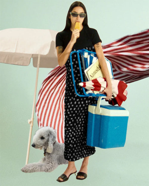 A person is wearing a black top and a long, black strapless dress with a sweetheart neckline and a halter neck strap. The dress features an all-over pattern of small, irregular white spots. The person is holding an ice cream cone and carrying a blue cooler, a beach chair, and a red and white striped umbrella. There is also a white dog lying on the ground and a white umbrella next to the person.