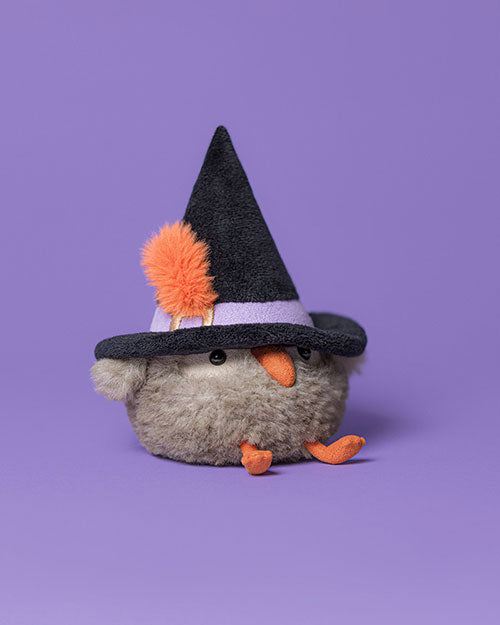 Grey plush bird toy with an orange beak and feet, wearing a black witch's hat with a purple band and orange pom-pom, set against a solid purple background.