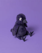 Plush toy bird with dark purple fur, large round eyes with black pupils and white sclera, a black beak, and outstretched legs, sitting on a purple surface.