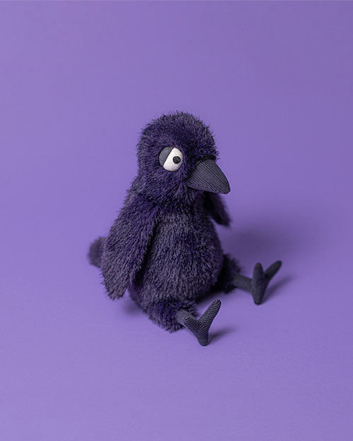 Plush toy bird with dark purple fur, large round eyes with black pupils and white sclera, a black beak, and outstretched legs, sitting on a purple surface.