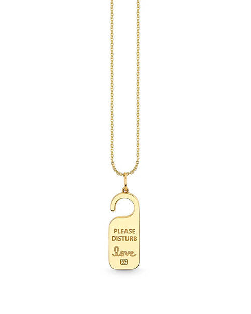 A gold necklace with a pendant designed to resemble a ‘Do Not Disturb’ hotel door hanger. The pendant has the phrase ‘PLEASE DISTURB’ engraved at the top and ‘Love’  below it.