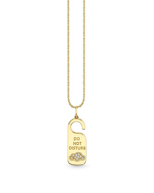 A gold necklace with a pendant designed to resemble a ‘Do Not Disturb’ door hanger. The pendant features an engraved message that reads ‘DO NOT DISTURB’ in capitalized, bold letters, with a cloud filled with pave diamonds at the bottom.