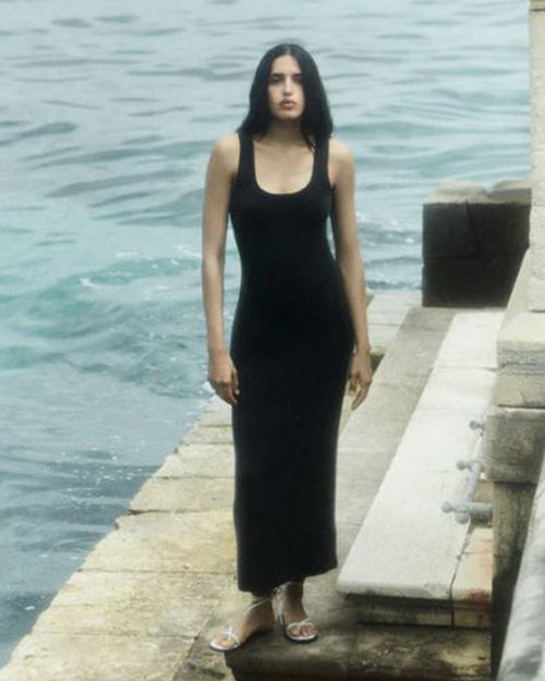 A person standing by a pier next to the water wearing a black maxi dress.