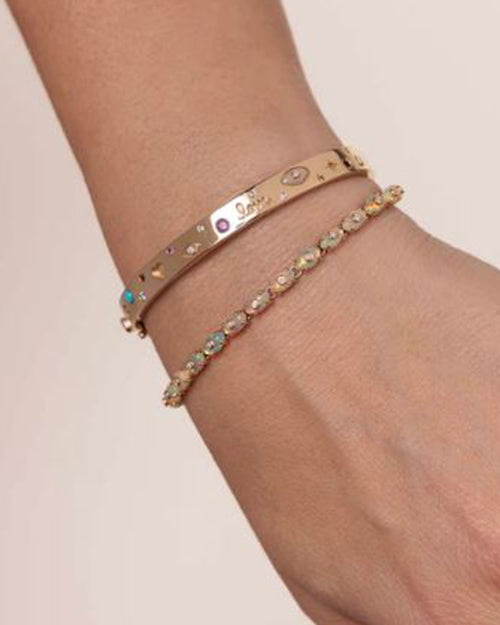 A gold bangle bracelet adorned with cut-out patterns. The word “Love” is engraved on the bracelet, which is further embellished with colorful gemstones of varying sizes and shapes. The bracelet is paired with a different colorful bracelet.