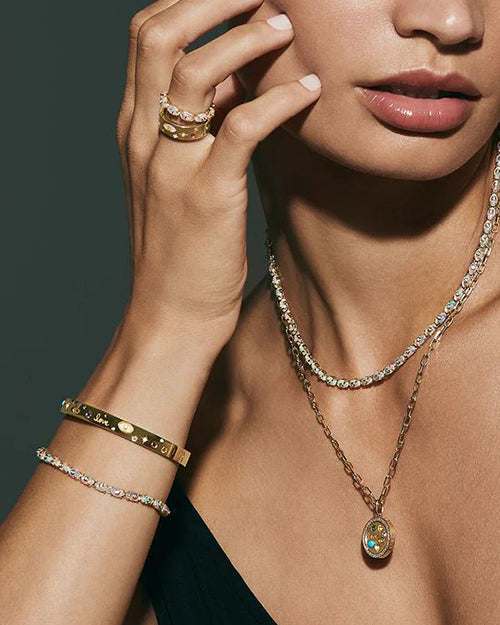 A model in a gold necklace featuring a circular pendant with the word ‘love’ embossed in the center, surrounded by small gemstones and diamonds. The chain is made of thick, interlocking gold links. Necklace is paired with matching bracelet and diamond chain.