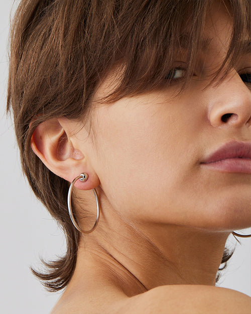 A close-up headshot of a brunette models' ear wearing a silver hoop earring with a semi-circle detail at the lobe. The background is plain white.