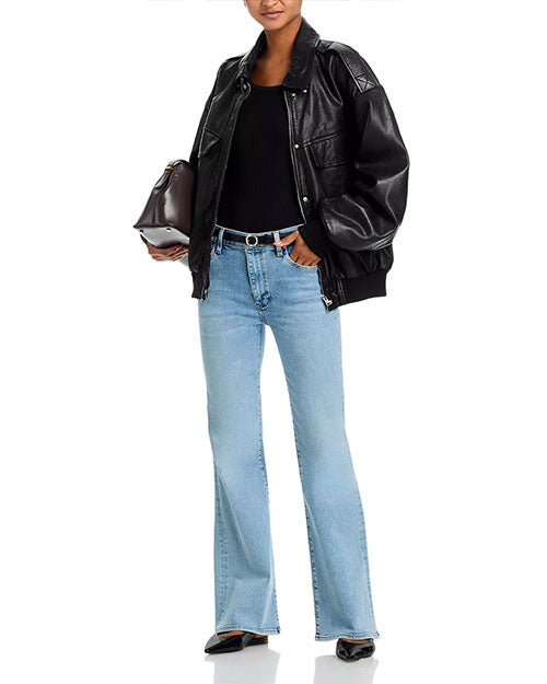 Person wearing an oversized black leather jacket over a black top, light blue flared jeans, black belt, and black pointed shoes, holding a dark brown handbag.