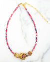 A beaded necklace featuring alternating pink and blue beads, with several gold rings and charms clustered at the center. The necklace has a gold chain extender attached at one end, displayed on a white marble surface.