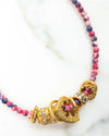 Close-up of a beaded necklace with alternating pink and blue beads, featuring a cluster of gold rings and charms at the center, including a heart-shaped piece and a decorative bead with engraved patterns.