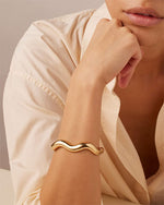 A woman in a white shirt wearing a gold bracelet, smiling softly at the camera.

