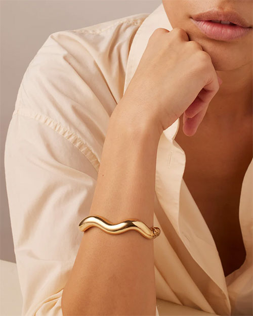 A woman in a white shirt wearing a gold bracelet, smiling softly at the camera.

