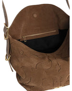 An open brown suede handbag with a decorative pattern, showing a black-lined interior with a zippered pocket. The bag features gold hardware, including buckles on the strap