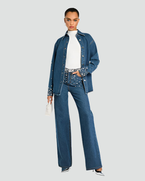 A full body shot of a brunette model standing in front of a white background, wearing high-waisted, wide-leg blue jeans with decorative rhinestone embellishments at the waistline to hips. The model is also wearing a long-sleeved white top tucked into the jeans, pointy heels, and a denim jacket with rhinestone embellishments at the wrist cuffs.