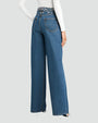 A backside view of the lower body of a model standing in front of a white background, wearing high-waisted, wide-leg blue jeans with decorative rhinestone embellishments at the waistline. The model is also wearing a long-sleeved white top tucked into the jeans with silver heels.