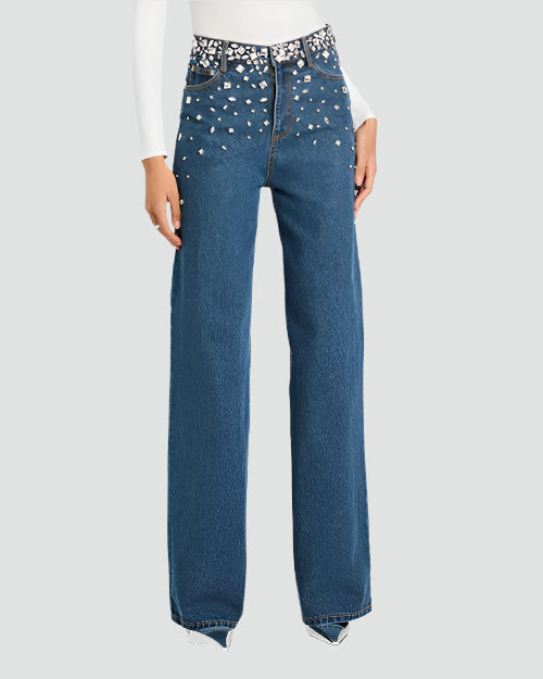 A lower body shot of a model standing in front of a white background, wearing high-waisted, wide-leg blue jeans with decorative rhinestone embellishments at the waistline to hips. The model is also wearing a long-sleeved white top tucked into the jeans, and pointy heels.
