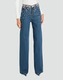 A lower body shot of a model standing in front of a white background, wearing high-waisted, wide-leg blue jeans with decorative rhinestone embellishments at the waistline to hips. The model is also wearing a long-sleeved white top tucked into the jeans, and pointy heels.