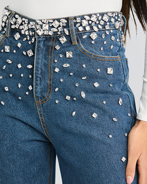 A close-up shot of the lower body of a model standing in front of a white background, wearing high-waisted, wide-leg blue jeans with decorative rhinestone embellishments at the waistline to hips. The model is also wearing a long-sleeved white top tucked into the jeans.