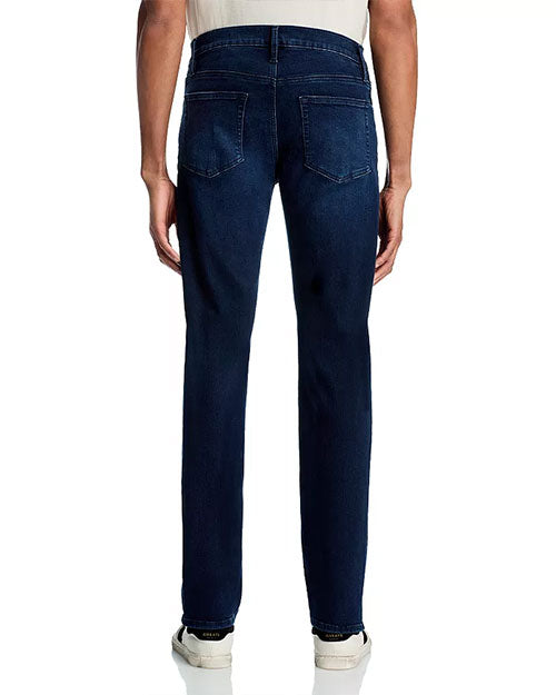 Back view of a person standing upright, wearing dark blue jeans with visible back pocket stitching and white sneakers. Only the lower torso and legs are visible.
