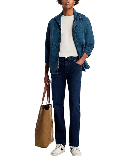 Person standing in a casual outfit: white t-shirt, blue denim jacket, dark blue jeans, white sneakers, and holding a large brown tote bag in the left hand.