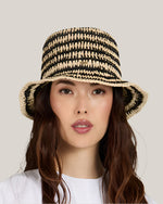 Front view of a woman wearing a black and beige striped straw hat  and a white shirt on a plain background