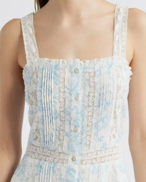 A close-up frontal image of a person wearing a light blue and white dress with a small blue floral pattern, buttoning down the front and over the shoulder straps. The dress has a square neckline and features vertical pleats along the front. There are lace details running vertically alongside the pleats.