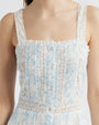 A close-up frontal image of a person wearing a light blue and white dress with a small blue floral pattern, buttoning down the front and over the shoulder straps. The dress has a square neckline and features vertical pleats along the front. There are lace details running vertically alongside the pleats.