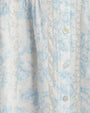 An extremely close-up view of the fabric on a light blue and white dress. The dress has a light blue floral pattern and is buttoned down the front with small buttons. The dress features vertical pleats along the front. There are lace details running vertically alongside the pleats