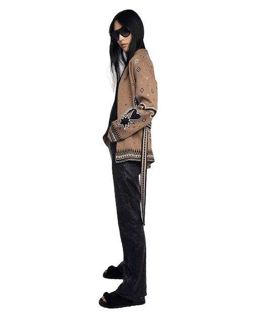 Person standing in profile view, wearing dark sunglasses, a beige jacket with black and white patterns, black pants, and black open-toe shoes. 