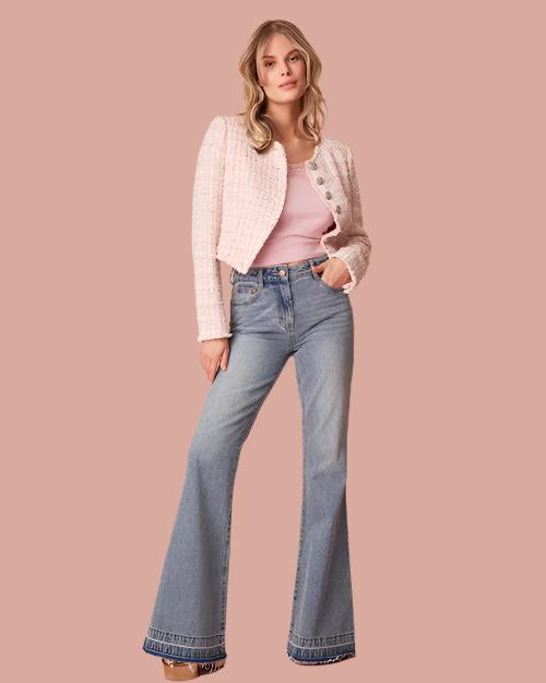 A full body frontal view of a blonde model with her hand in her pocket wearing a pair of light blue bootcut jeans with a double ankle hem. The model is pairing the jeans with a white button-down sweater over a pink top. The background is plain pink.