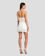 A full body backside view of a brunette model standing against a plain grey background and wearing a short white dress with intricate geometric and floral details at the hem and waist, as well as an open back design. The model is accessorized with large round earrings, a gold bracelet and white high-heeled sandals.