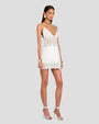 An angled full-body view of a brunette model standing against a plain grey background and wearing a short white dress with intricate geometric and floral details at the hem, waist and neckline. The model is accessorized with large round earrings, a gold choker necklace, and thick gold bracelets.