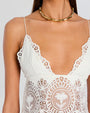A close-up chest-level shot of a model wearing a white lace dress with intricate geometric and floral patterns around the waist and the thin strapped V-neck, with a scalloped design. The model is also wearing an old bamboo designed choker necklace.