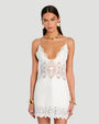 A knee-up shot of a brunette model standing against a plain grey background and wearing a short white dress with intricate geometric and floral details at the hem, waist and neckline. The model is accessorized with large round earrings, a gold choker necklace, and thick gold bracelets.