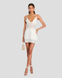 A full body shot of a brunette model with her legs crossed and her hands on her hips, standing against a plain grey background and wearing a short white dress with intricate geometric and floral details at the hem, waist and neckline. The model is accessorized with large round earrings, a gold choker necklace, gold bracelets, and white high-heeled sandals with a beige handbag.