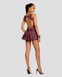 A full body backside view of a brunette model with her hair up and her hand on her hip, wearing a sleeveless dress a diamond open-back design and a maroon and black leaf pattern, featuring a fitted bodice and a flared skirt with a black belt. She is also wearing gold hoop earrings, bangles, and black open-toed heels. The background is plain grey.