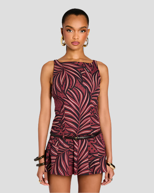 A knee-up view of a brunette model with her legs crossed wearing a sleeveless dress with a maroon and black leaf pattern, featuring a fitted bodice and a flared skirt with a black belt. She is also wearing gold hoop earrings and black and gold bangles.
